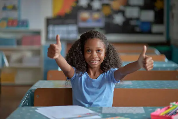 Portrait of a happy young girl giving thumbs up while sitting at her desk in a classroom