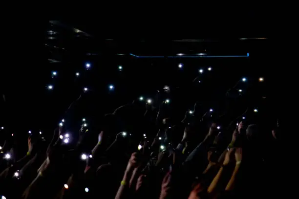Photo of Group of people holding cigarette lighters and mobile phones at a concert crowd of people fans with their hands up. Bright lights.