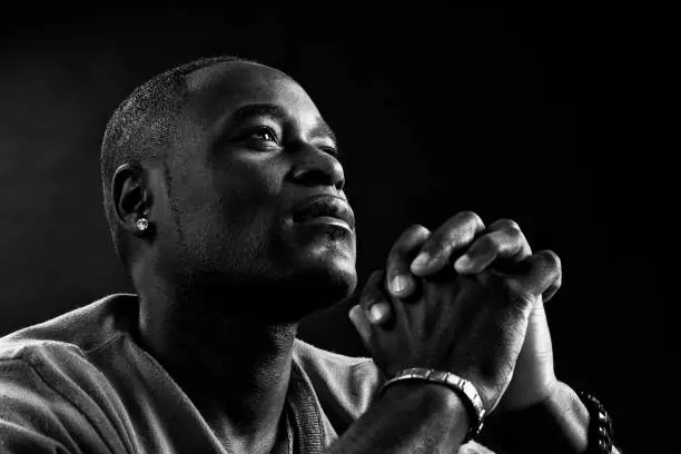 A black man, African American or of African descent, looks into the distance, his hands clasped, praying, in this black and white portrait.
