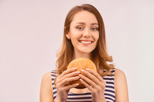 Happy pretty girl gently smiles snow-white beautiful smile, wears shiny earring in ear, striped t-shirt, eats delicious Burger, fast food, isolated on white background in Studio with empty space text