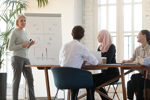 Middle-aged businesswoman stand lead meeting presenting business financial project on whiteboard to multiracial employees, mature female speaker make presentation for diverse colleagues at briefing