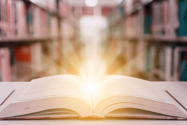 istock Rays of light coming from an open book or textbook on wooden table with blurred bookshelf in the library. Success education learning or back to school concept background 1205483523