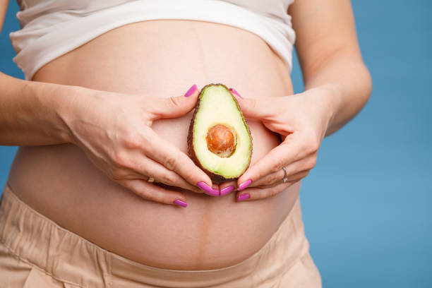 Pregnant woman holding half of avocado next to her belly stock photo