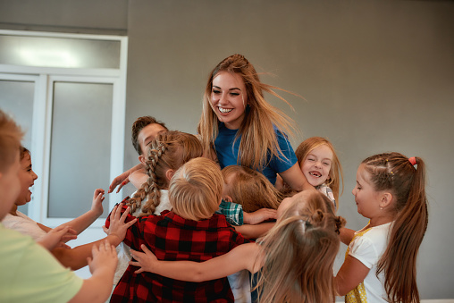 Young positive female dance teacher hugging happy children and smiling while standing in studio. Relationship between teacher and kids. Choreography class. Dance school
