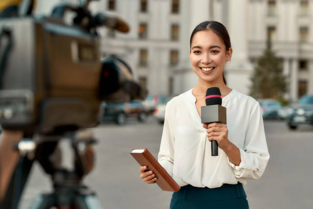 On air. TV reporter presenting the news outdoors. Journalism industry, live streaming concept. Portrait of professional female reporter at work. Young asian woman standing on the street with a microphone in hand and smiling at camera. Horizontal shot. Selective focus on woman journalism stock pictures, royalty-free photos & images