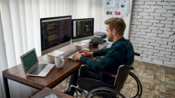 istock Professional programmer. Side view of young concentrated web developer in a wheelchair writing program code on multiple computer screens while sitting at his workplace in the modern office 1205489097