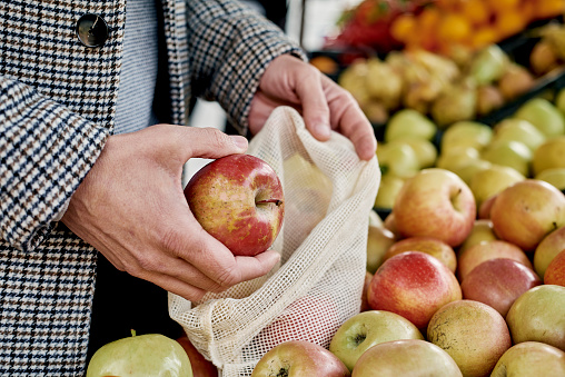 closeup of a man shopping at a greengrocer putting some red apples in a textile reusable mesh bag, as a measure to reduce plastic pollution