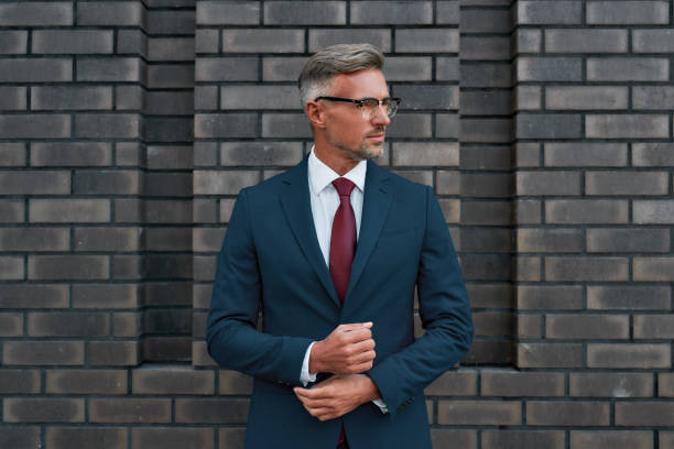istock Confident and stylish. Handsome mature businessman in full suit adjusting his sleeve while standing outdoors against the brick wall 1205489843