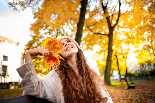 Outdoor lifestyle portrait of young woman with warm hat holding colorful autumn leaves and smiling