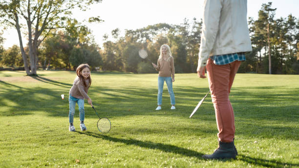 rozwijanie się poprzez zabawę i kreatywność. rodzice bawią się z córką w badmintonie w parku w słoneczny dzień - child laughing blond hair three people zdjęcia i obrazy z banku zdjęć