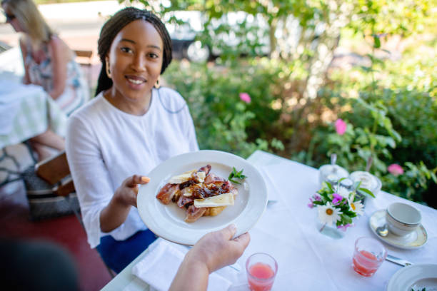 Personal perspective of a waiter serving a breakfast to a friendly guest at a cafe restaurant on an outdoor porch veranda.  There is guava juice on the table and flowers and a cup and saucer.  The girl is being served breakfast.  She is an attractive beautiful African woman in her twenties.