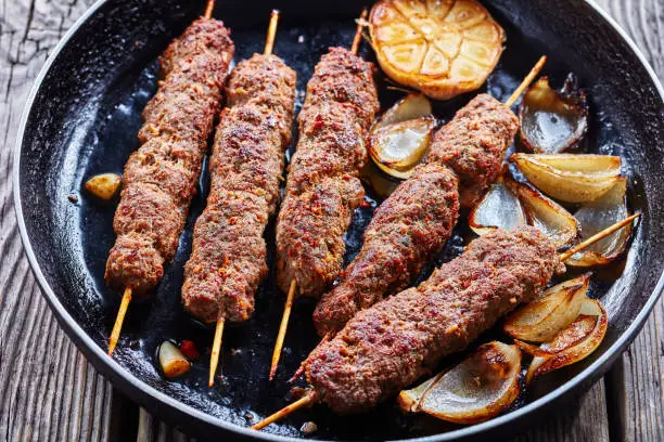 Traditional middle east kefta or kofta kebab of a mix of ground beef and lamb meat grilled on skewers served on a skillet with roasted garlic and onion on a table of barn wood, close-up