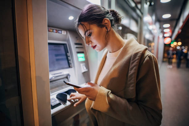 Young teenage girl using ATM machine and smart phone One young beautiful young teenage girl using ATM machine and smart phone in the city atm photos stock pictures, royalty-free photos & images