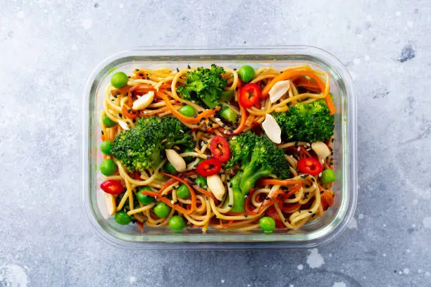 Stir fry noodles, udon with vegetables in glass lunch box. Grey stone background. Top view.