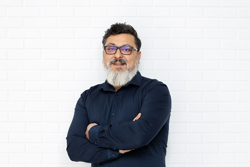 Relaxed middle eastern senior business man with glasses and stylish beard smiling on brick wall, Portrait of successful multi-ethnic trendy old bearded entrepreneur with crossed arms feeling satisfied