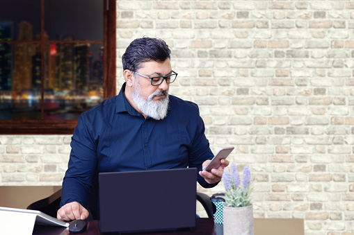 Successful mature middle eastern businessman wearing eyeglasses working on laptop while using phone in modern office – Serious indian senior man entrepreneur sitting at desk working late