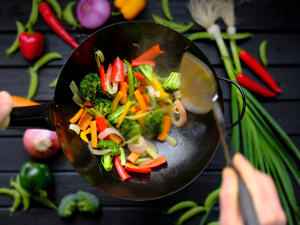 Stir frying and sauteing fresh colorful market vegetables in a hot wok. A variety of fresh market vegetables including broccoli, carrot, capsicum, red onion, green beans, and scallion being stir-fried and cooked in a hot wok with fresh vegetables on a dark black wooden table in the background. sauteed stock pictures, royalty-free photos & images