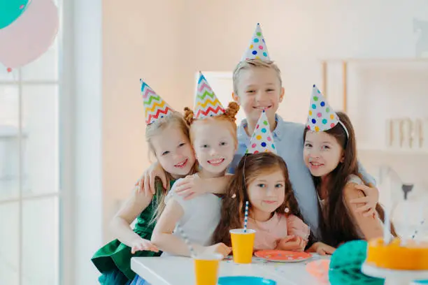 Photo of Photo of happy friends gather around table, wear party hats, embrace and look gladfully at camera, smile positively, celebrate birthday at home together, have fun