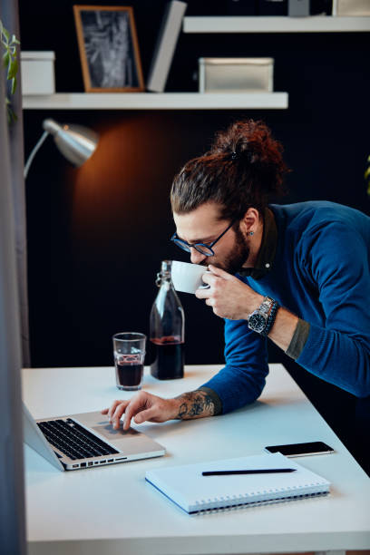 istock Young attractive bearded hipster with curly hair leaning on table, drinking coffee and surfing on internet while standing in home office. 1205509041