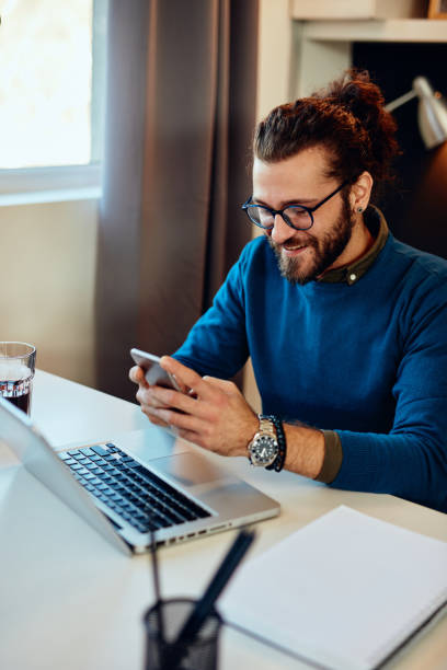 istock Smiling caucasian hipster sitting in his office and using smart phone for responding an e-mail. 1205509238