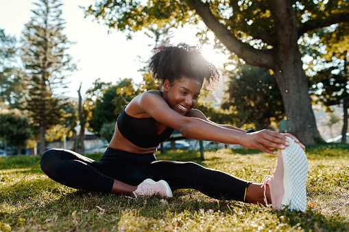 Retrato sonriente de una joven americana afroamericana de ajuste deportivo sentada en el césped estirando las piernas en el parque - joven mujer negra feliz calentando sus muslces antes de correr photo