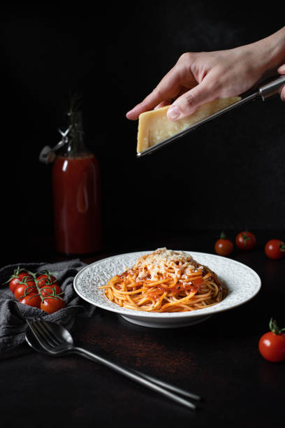 istock Pasta with tomato sauce and parmesan. White plate with spaghetti on a dark background. Hands grating Parmesan cheese on a spaghetti pasta. Traditional Italian food. Low key, vertical, copy space 1205534980