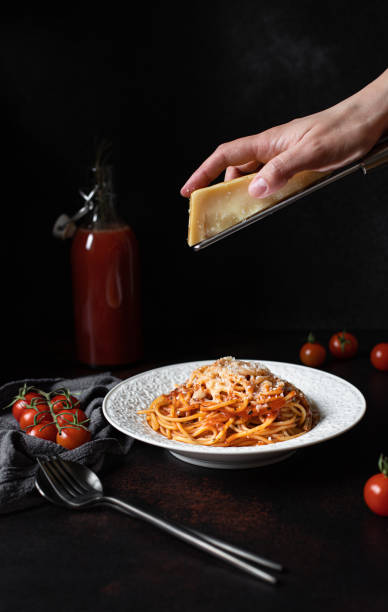 istock Pasta with tomato sauce and parmesan. White plate with spaghetti on a dark background. Hands grating Parmesan cheese on a spaghetti pasta. Traditional Italian food. Low key, vertical, copy space 1205536239