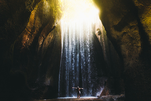 Young woman feeling awe and happiness getting wet staying under the huge waterfall hidden in the cave during bright summer sunny sunset on Bali, Indonesia