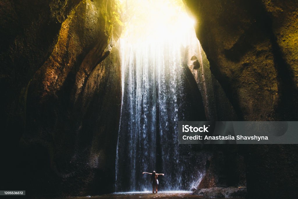 Woman meets sunrise in the cave under the big waterfall on Bali island, Indonesia Young woman feeling awe and happiness getting wet staying under the huge waterfall hidden in the cave during bright summer sunny sunset on Bali, Indonesia Waterfall Stock Photo