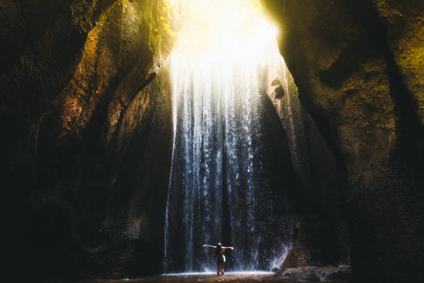 Woman meets sunrise in the cave under the big waterfall on Bali island, Indonesia Young woman feeling awe and happiness getting wet staying under the huge waterfall hidden in the cave during bright summer sunny sunset on Bali, Indonesia falling water flowing water stock pictures, royalty-free photos & images