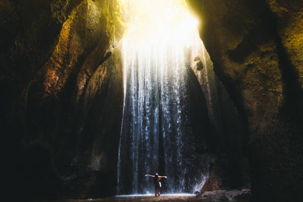 Woman meets sunrise in the cave under the big waterfall on Bali island, Indonesia Young woman feeling awe and happiness getting wet staying under the huge waterfall hidden in the cave during bright summer sunny sunset on Bali, Indonesia cataract photos stock pictures, royalty-free photos & images