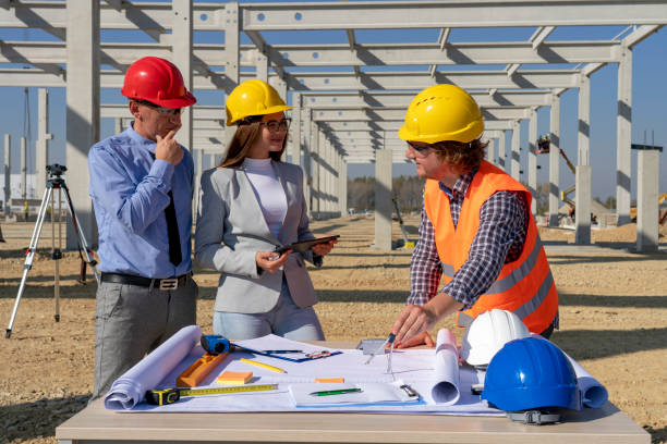 istock Mixed Construction Team in Yellow and Red Hardhats Discuss on Blueprint on Site Under Construction 1205538083