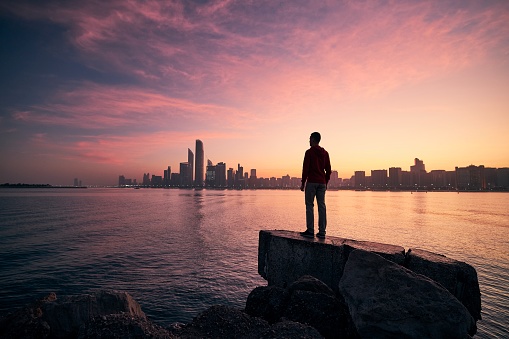 Young man standing on seashore and watching colorful sunrise. Sea and urban skyline Abu Dhabi, United Arab Emirates.