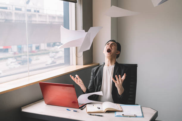 un jeune ouvrier blanc chinois asiatique jetant des papiers dans l’air frustré et le stress émotif dans son bureau s’asseyant sur sa chaise - frustration computer men emotional stress photos et images de collection