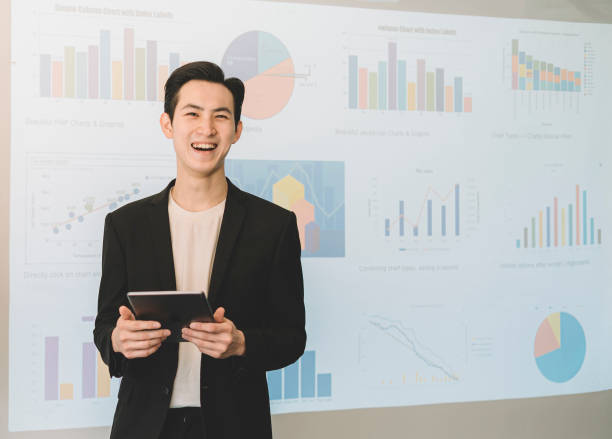 istock an asian chinese young white collar worker standing in front of the projector screen with all forecast and projection chart laughing and holding a digital tablet 1205543863