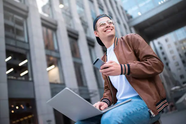 Photo of Smart attitude. Positive handsome man using a laptop and sitting in the street while surfing the internet have a web conference. Web chat online conference.