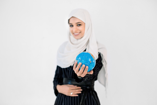 Portrait of a happy Arabian muslim woman in hijab holding world globe isolated on a white background. Focus on the globe.