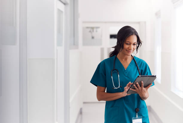 female doctor wearing scrubs in hospital corridor using digital tablet - health imagens e fotografias de stock