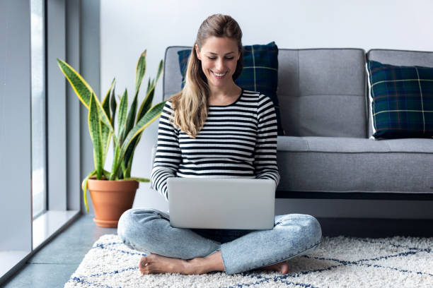 pretty young woman using her laptop while sitting on the floor at home. - furniture internet adult blond hair imagens e fotografias de stock