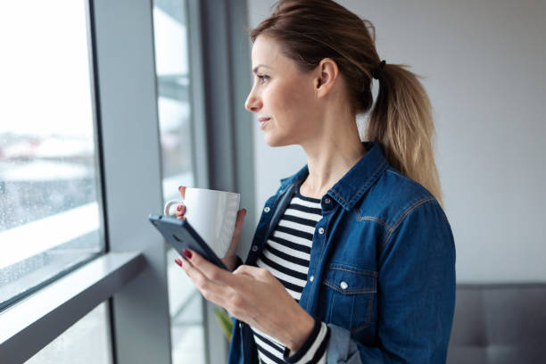 istock Pretty young woman using her mobile phone while drinking coffee near to the window in the living room at home. 1205573430