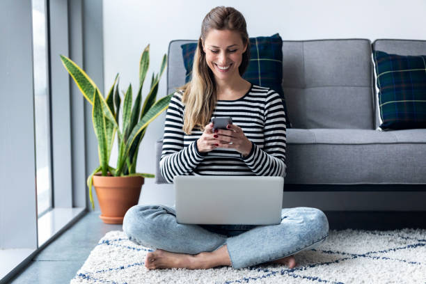 pretty young woman using her mobile phone while working with laptop sitting on the floor at home. - furniture internet adult blond hair imagens e fotografias de stock