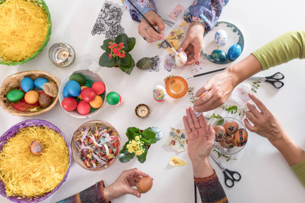 istock White table with colored Easter eggs in baskets and bowls on it, along with a lot of arts and crafts tools. Three women are painting some of the eggs by hand using decoupage technique. 1205576771
