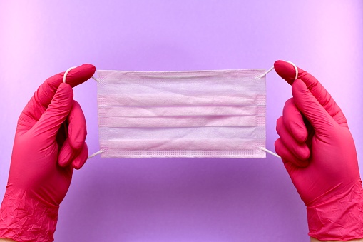 The mask is medical purple stretched behind rubber bands fastening hands in rubber fuchsia-colored rubber gloves in the center on a purple background with light spots.