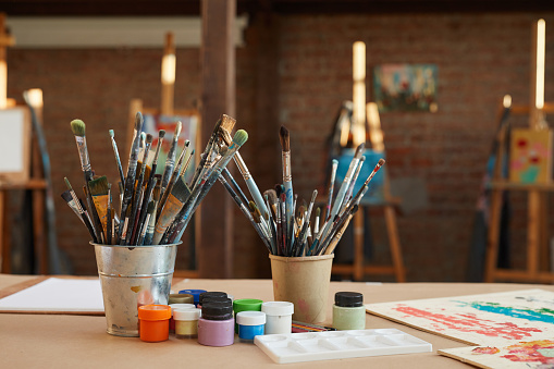 Image of different kinds of brushes with colored paints and pictures are on the table in workshop