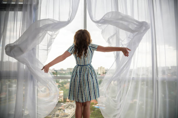 vista posteriore di una giovane donna gioiosa che indossa un abito di moda e tiene le tende aperte per guardare fuori dalla grande finestra luminosa di casa, rivolgendosi a guardare e sorridere alla fotocamera, agli interni - curtain window women opening foto e immagini stock