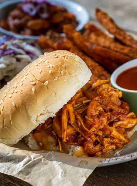 Vegan BBQ sandwich made with jack-fruit accompanied by baked beans, roasted sweet potato fries and coleslaw.