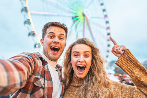 Happy couple having fun at amusement park in London - young couple in love taking a selfie and enjoying time at funfair with rollercoaster on background - Happy lifestyle and love concepts
