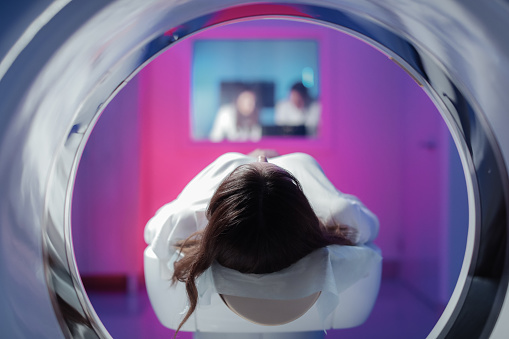 The girl patient is lying in the tomograph and waiting for a scan. Three doctors from the exam room look at the pictures.