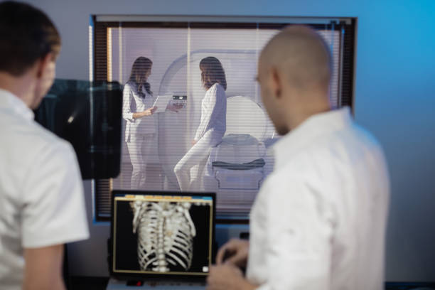 In Control Room Doctor and Radiologist Discuss Diagnosis while Watching Procedure, In the Background Patient Undergoes MRI or CT Scan Procedure. stock photo