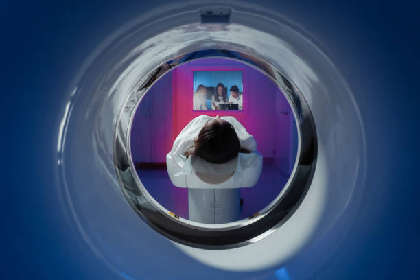 The girl patient is lying in the tomograph and waiting for a scan. Three doctors from the exam room look at the pictures stock photo